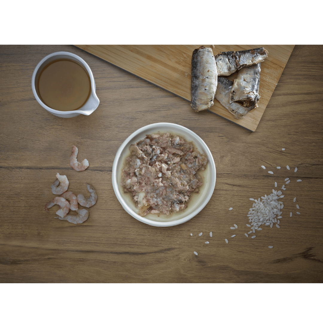 Some fresh ingredients, sardine, shrimp, broth and rice. In the middle of these, there is a bowl of Fish 4 Cats Sardine & Shrimp served in a bowl.
