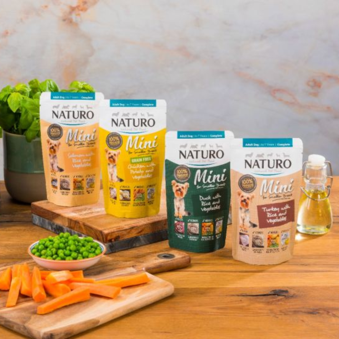 4 naturo dog food pouches on a wooden counter, surrounded by raw ingredients.