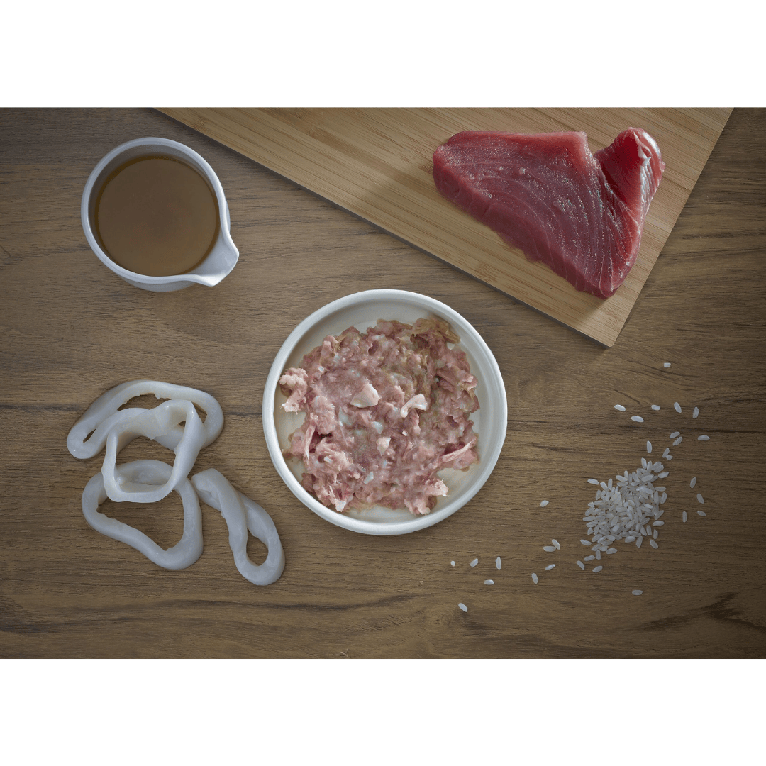 Raw Tuna, squid rings, and rice on a wooden surface with a small bowl of broth. There is a bowl of Fish 4 Cats Tuna with Squid in the middle
